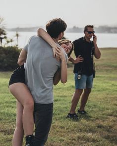 a man and woman hug each other as they stand in the grass near a body of water