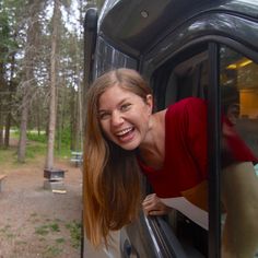 a smiling woman leaning out the window of a vehicle in front of a camper