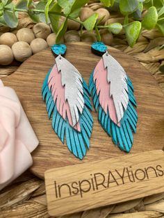 two pairs of pink and blue feather earrings sitting on top of a wooden board next to rocks