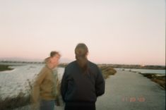 two people walking down a path near the ocean at sunset or dawn with one person looking on