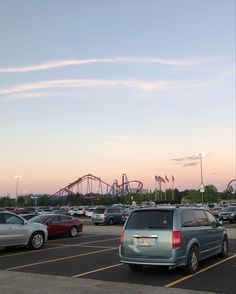 several cars parked in a parking lot with roller coasters in the background at dusk