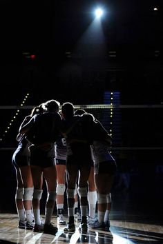 a group of women in shorts huddle together on a court at night with their backs turned to the camera