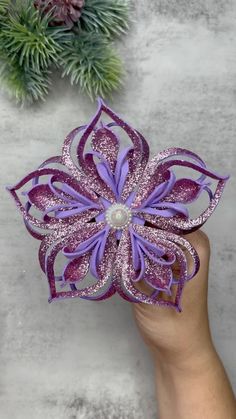 a hand holding a purple and white flower on top of a gray table next to pine cones