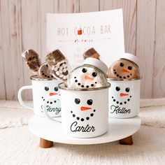 three mugs with snowmen in them sitting on a white plate next to a sign that says hot cocoa bar