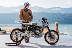 a man standing next to a motorcycle on top of a wooden platform near water and mountains