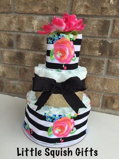 a three tiered diaper cake with flowers and black ribbon on top, sitting in front of a brick wall