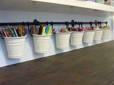 several white buckets with pens and pencils in them on a shelf above a counter