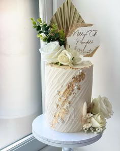 a wedding cake with white flowers and greenery sits on a table in front of a window