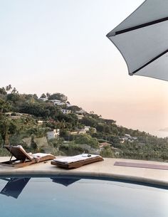 two lounge chairs and an umbrella on the edge of a swimming pool overlooking a hillside