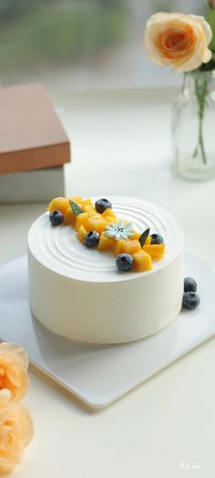 a white cake with blueberries and oranges on it sitting on a plate next to flowers