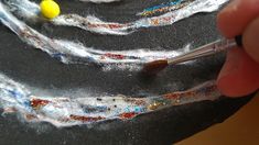 a hand holding a paintbrush and painting some white yarn on a black surface with yellow balls in the background