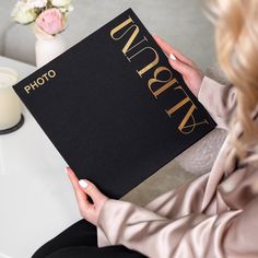 a woman sitting at a table holding up a black book with gold lettering on it