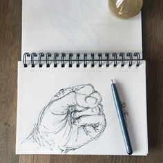 a drawing of a hand holding a ballpoint pen next to a spiral notebook on a wooden table