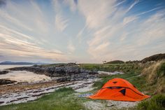 an orange tent sitting on top of a lush green field next to the ocean under a cloudy blue sky