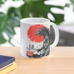 a coffee mug with godzilla on it sitting on a table next to books and a plant