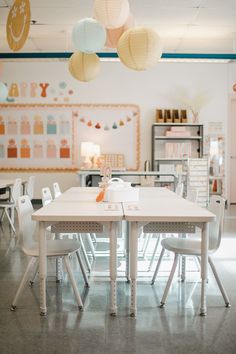 an empty classroom with white tables and chairs, paper lanterns hanging from the ceiling over them