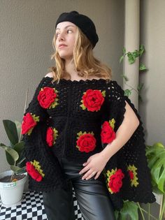 a woman wearing black and red crocheted sweater with roses on the shoulders, standing in front of a potted plant