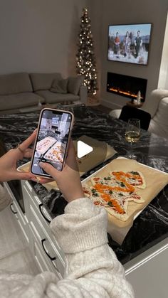 a woman taking a photo of pizza on her phone while sitting in front of the fireplace