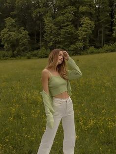 a woman standing in a field wearing white pants and a green crop top with one hand on her head