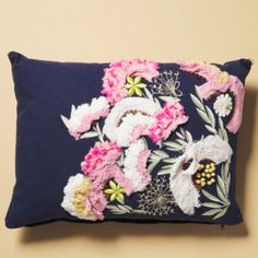 a blue pillow with pink and white flowers on the front, sitting against a tan wall