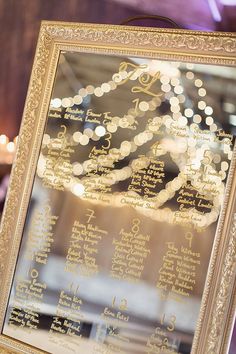 a wedding seating chart is displayed in a gold framed frame with string lights behind it