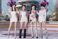 four women in short dresses and high heeled boots standing on a stage with flowers behind them