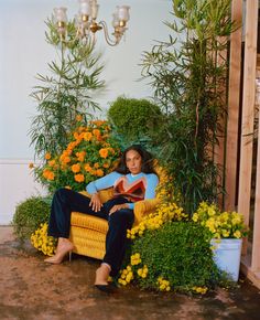 a woman sitting on a chair in front of some flowers and potted plants, reading a book