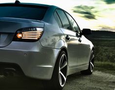 the front end of a silver car parked on a dirt road with grass and clouds in the background