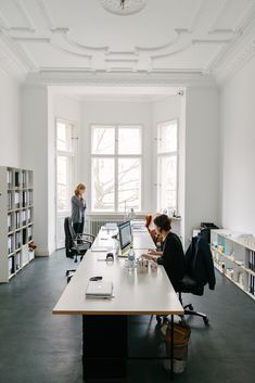 two people sitting at a desk in an office with bookshelves and open laptops