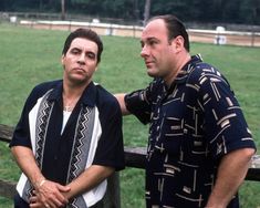 two men standing next to each other in front of a wooden fence and grass field