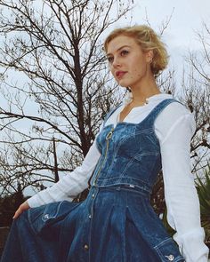 a woman sitting on top of a wooden bench wearing a jean dress and white shirt