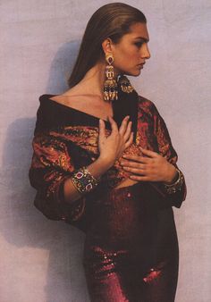 a woman with her hands on her chest in front of a white wall wearing jewelry