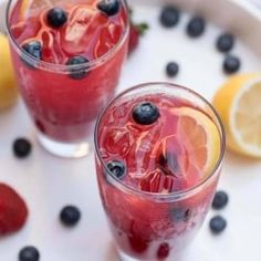 two glasses filled with fruit and ice on a plate next to lemons, strawberries, and blueberries