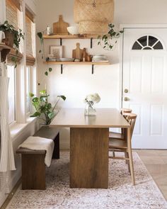 a dining room table and bench with plants on the shelf above it in front of a white door