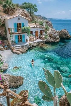 a person swimming in the water next to a house and some cactus plants on the shore