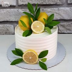 a white cake with lemons and green leaves on top, sitting in front of a brick wall