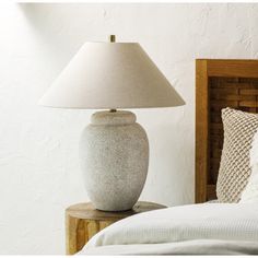 a white lamp sitting on top of a wooden night stand next to a bed with pillows