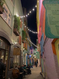 people are sitting on benches in an alleyway with lights strung above the buildings and below them