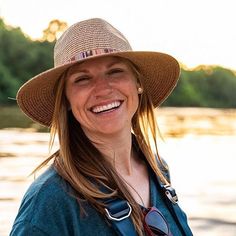 a woman wearing a hat and sunglasses standing next to a body of water with trees in the background