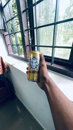 a hand holding a can of beer next to a window