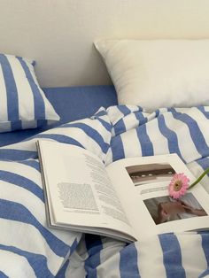 an open book sitting on top of a bed next to pillows and a pink flower