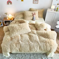 a bed covered in fluffy white blankets and pillows next to a night stand with stuffed animals on it