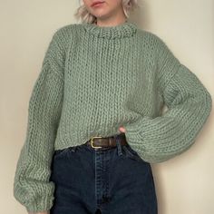 a woman wearing a green sweater and jeans standing in front of a wall with her hands on her hips