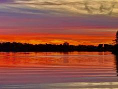 an orange and pink sunset over water with trees in the foreground, and clouds in the background