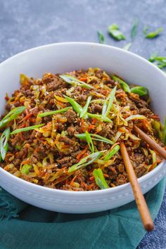 a white bowl filled with meat and vegetables next to chopsticks on a blue cloth