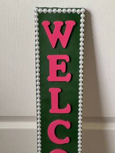 a welcome sign with the word welcome on it in pink and green colors, hanging against a white wall