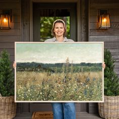 a woman holding up a large painting in front of a house with potted plants