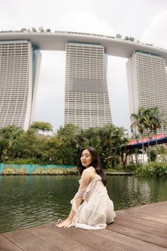 a woman in a white dress is sitting on a dock by some water and buildings