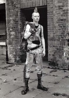 a man with punk hair standing in front of a brick building wearing boots and a vest