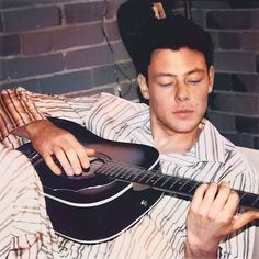 a man holding a guitar in his right hand while sitting on a couch next to a brick wall